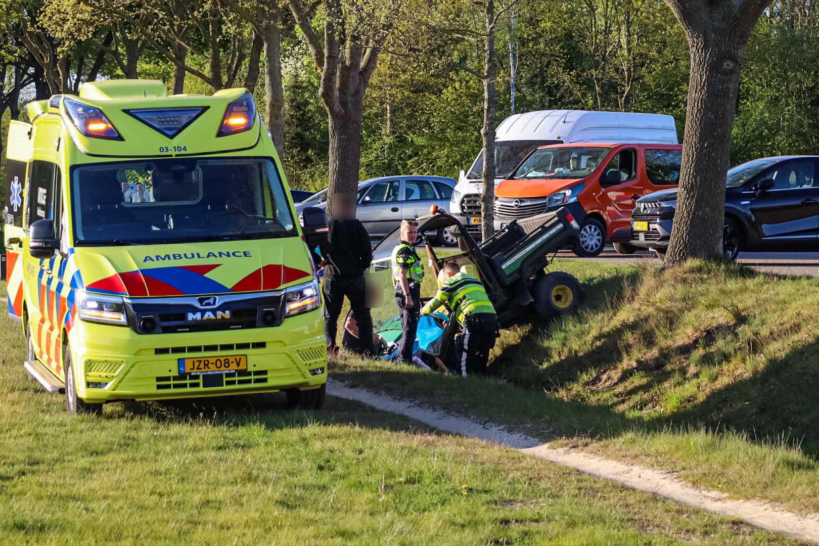 Bright yellow ambulance at a roadside as responders help a person from a ditch beside a toppled green vehicle on a sunny day.