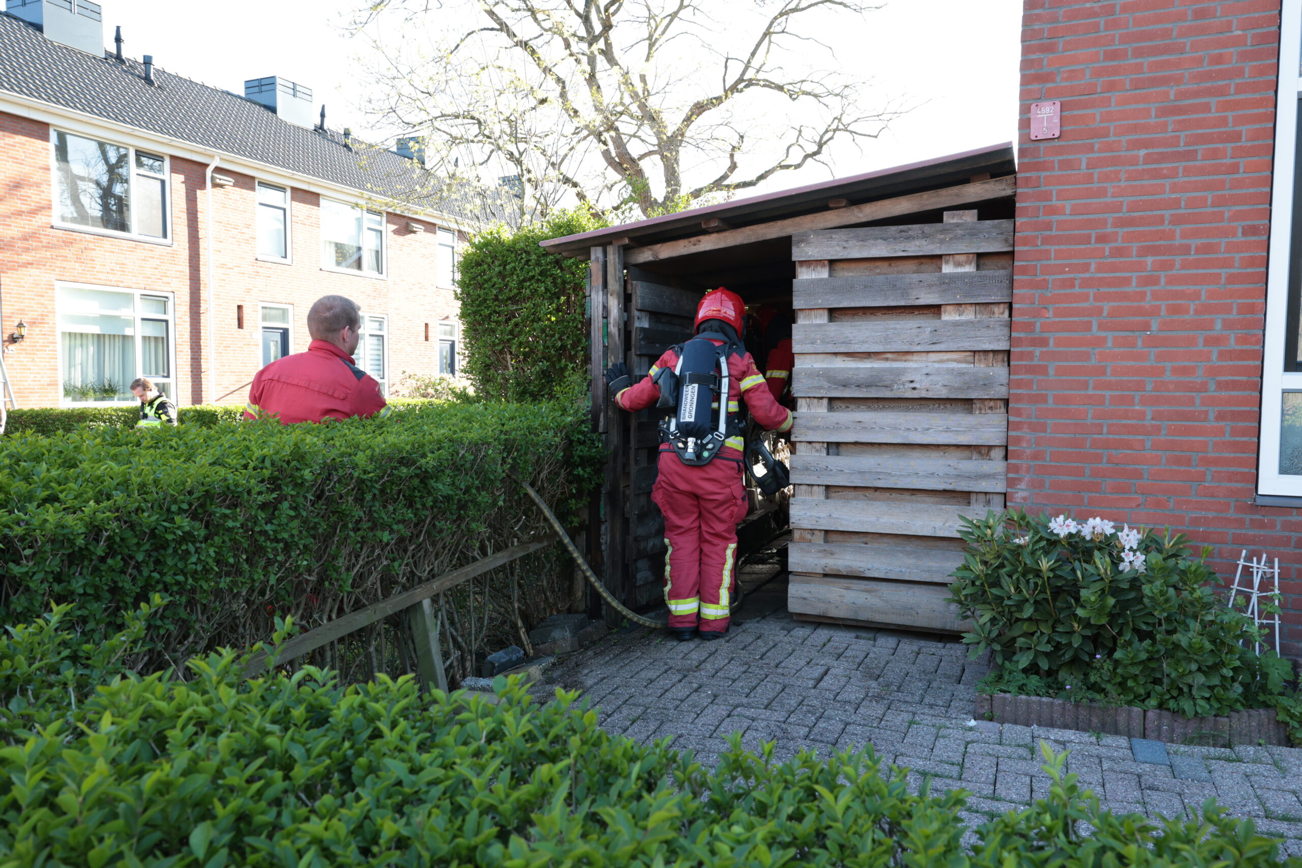 Firefighters in red gear entering a wooden shed or storage structure in a residential area, with brick houses and green hedges nearby.