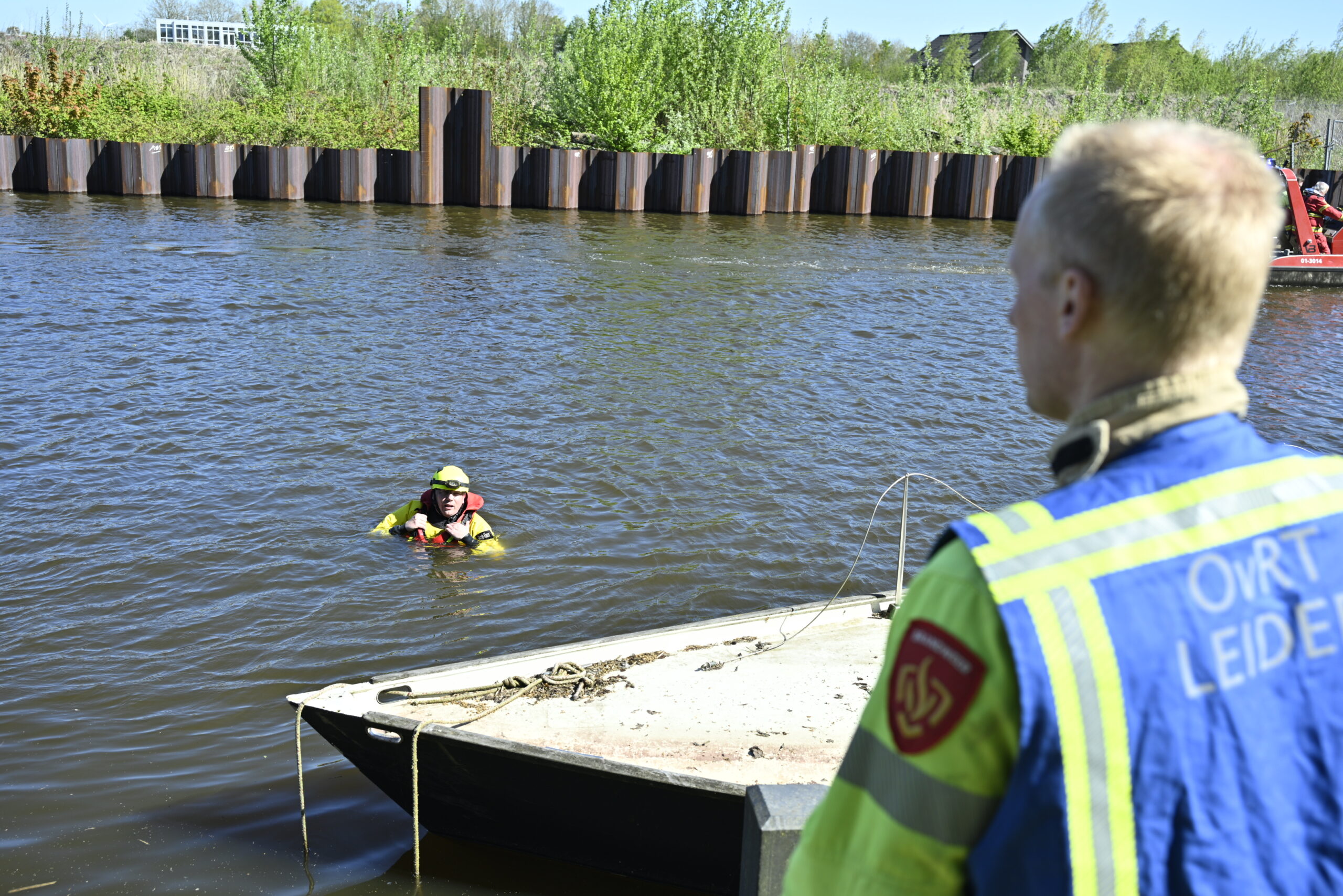 Two divers in yellow suits with red life jackets in the river near a small dock; a worker in a high‑visibility vest watches from shore in the foreground.