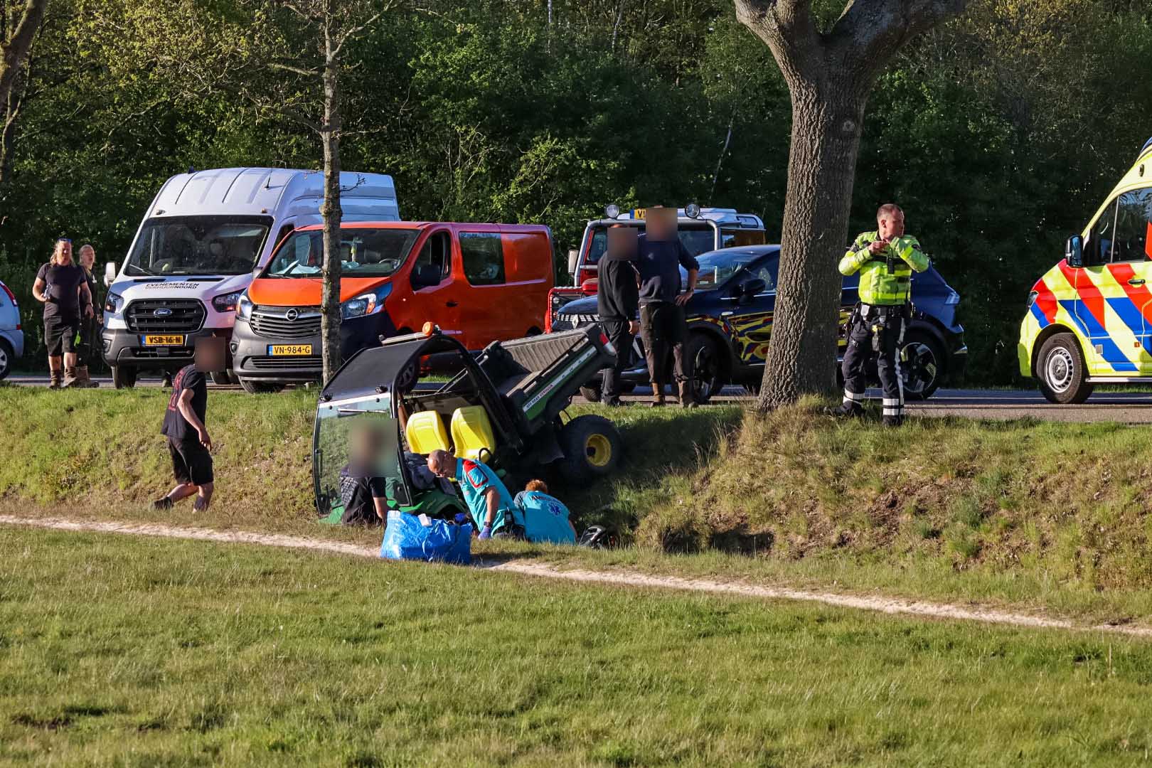 Roadside emergency scene with an overturned green tractor and paramedics assisting an injured person; multiple ambulances and police/response vehicles parked along a grassy verge in the background.
