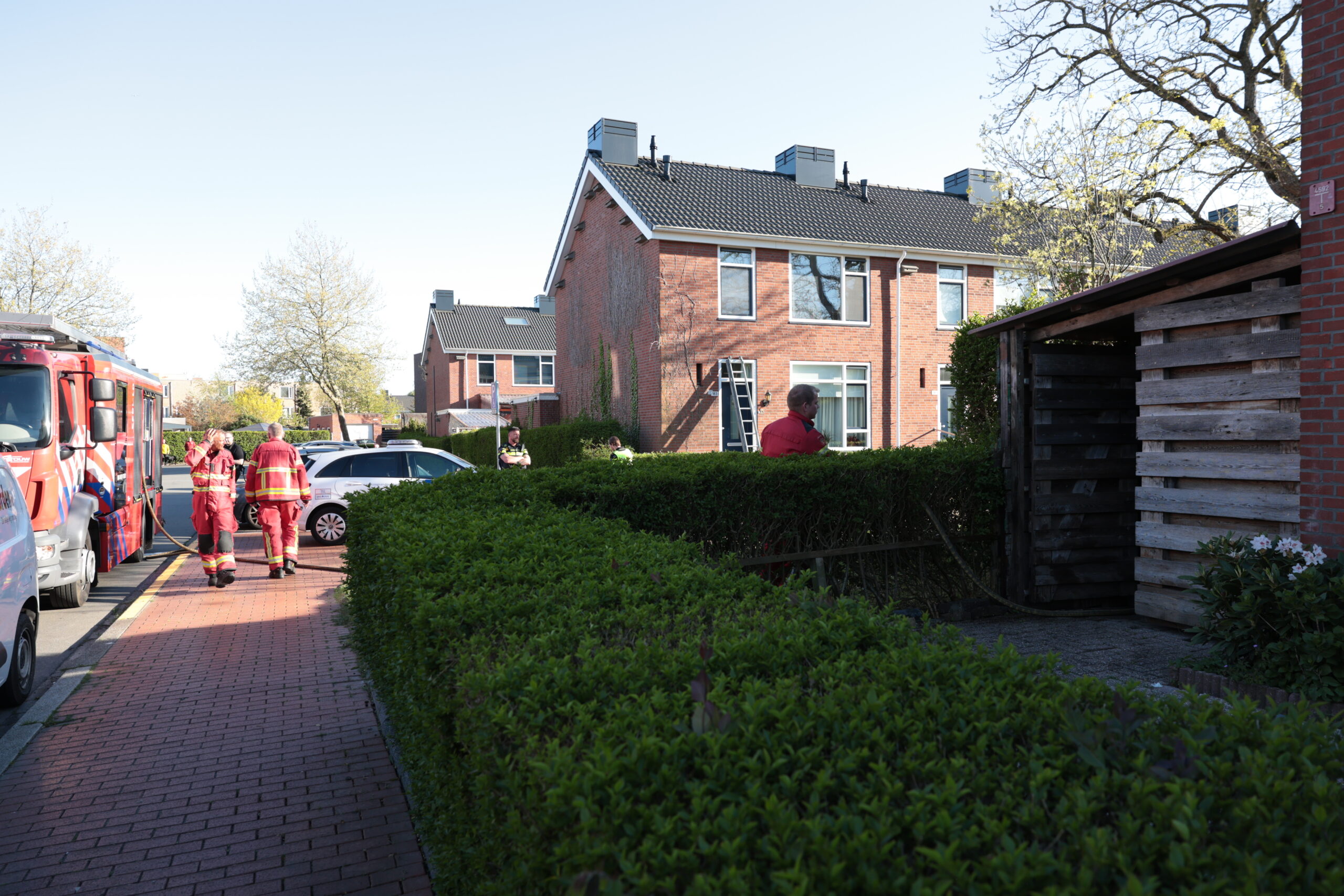 Firefighters in red turnout gear stand beside a red fire truck on a brick residential street during a daytime emergency.