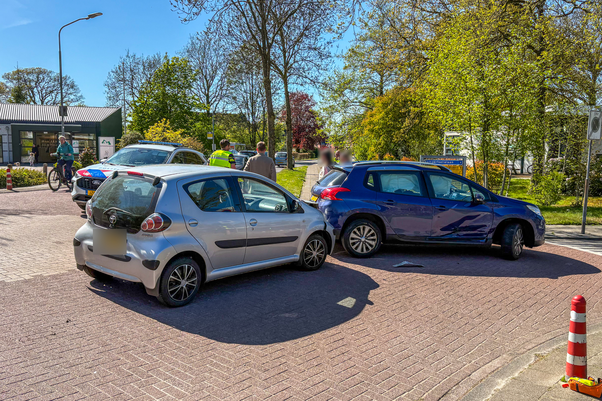 Street scene with multiple parked cars, a cyclist, and people near a storefront on a sunny day.