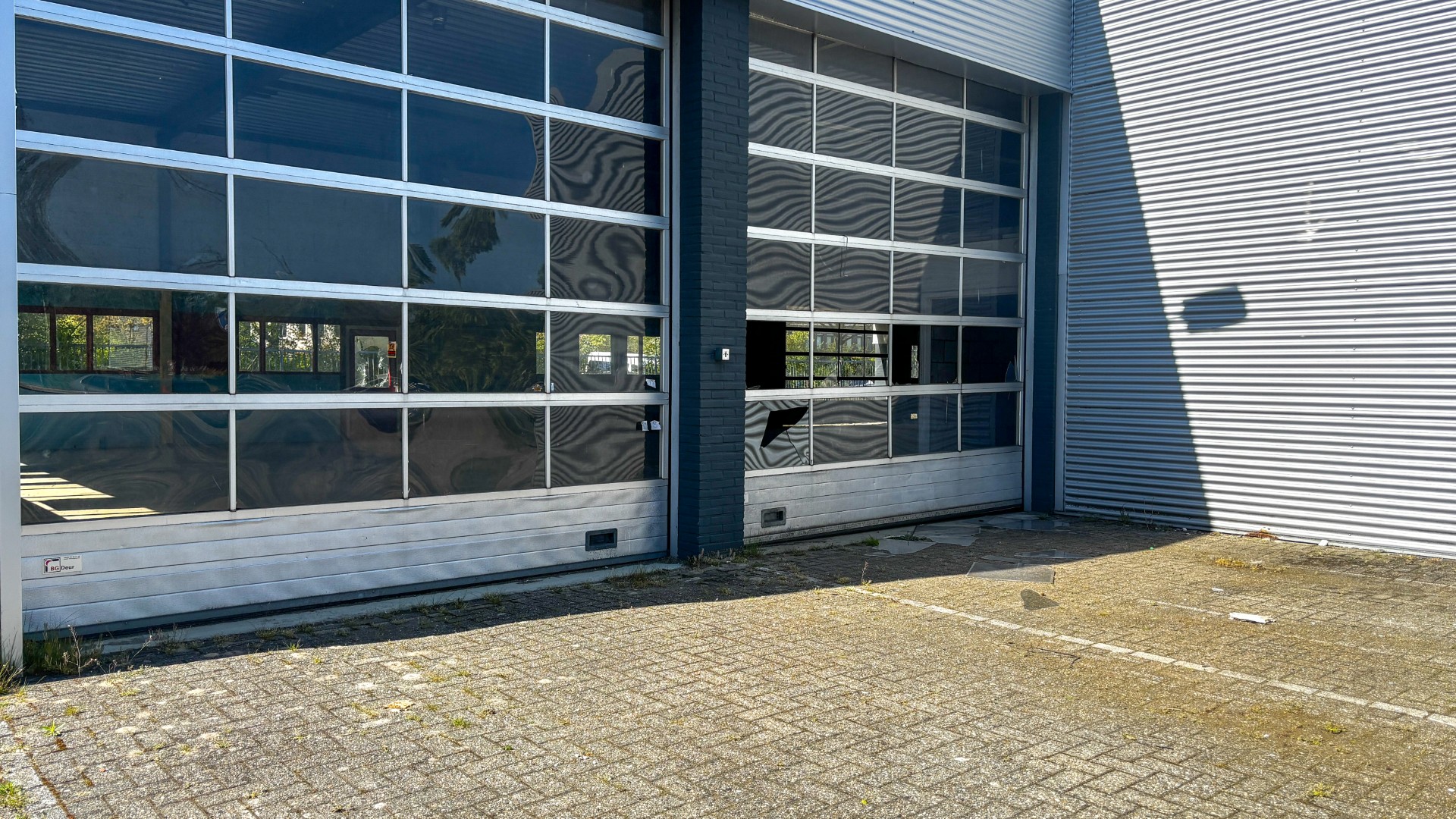 Industrial building facade with large segmented glass roll-up doors and corrugated metal wall; empty, sunlit parking area in foreground.