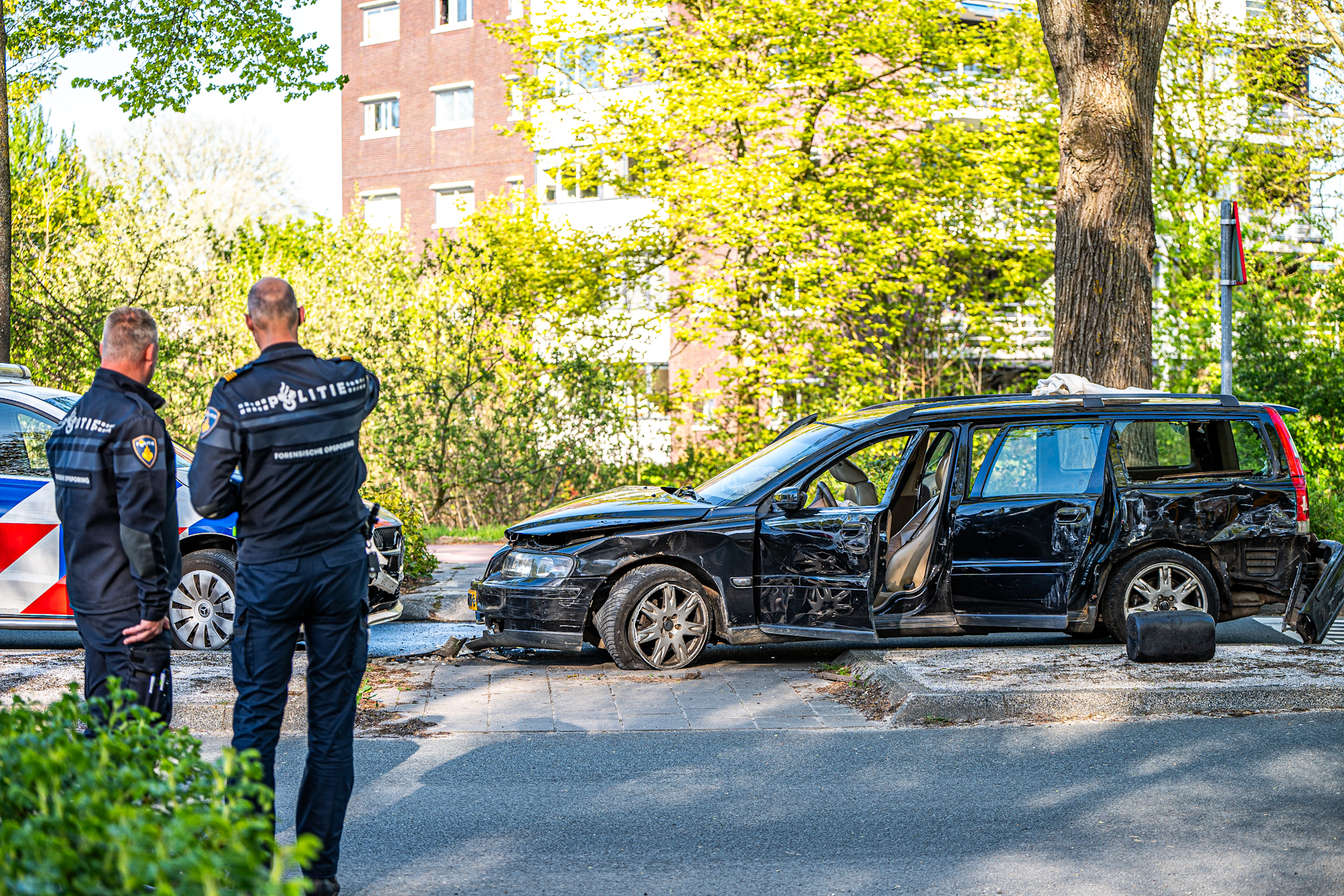 Two police officers in dark uniforms stand near a heavily damaged black SUV on a sunny street, with a police car and green trees in the background.