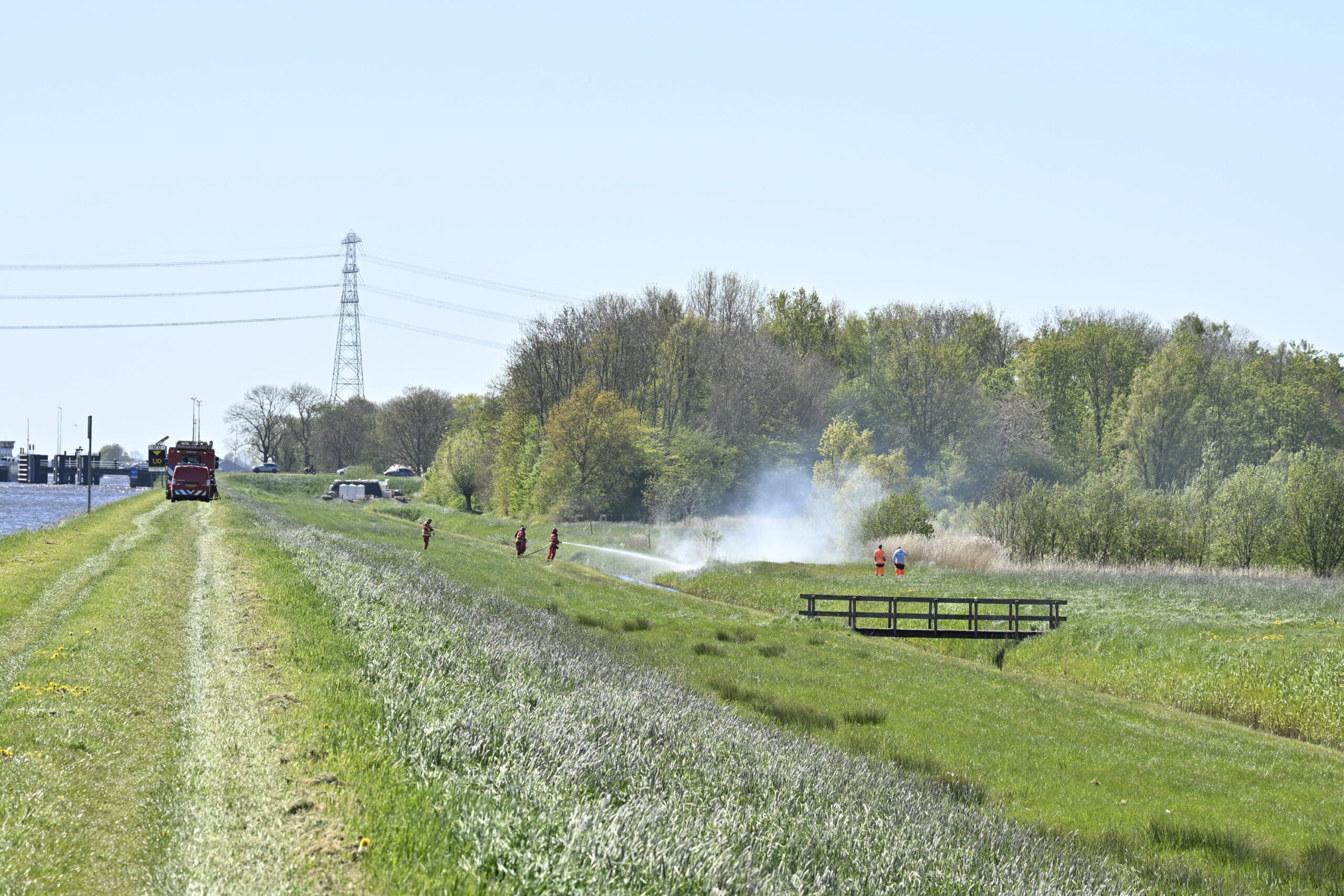 Firefighters in red gear spray water on a brush fire in a grassy field, with trees and a power tower in the distance.