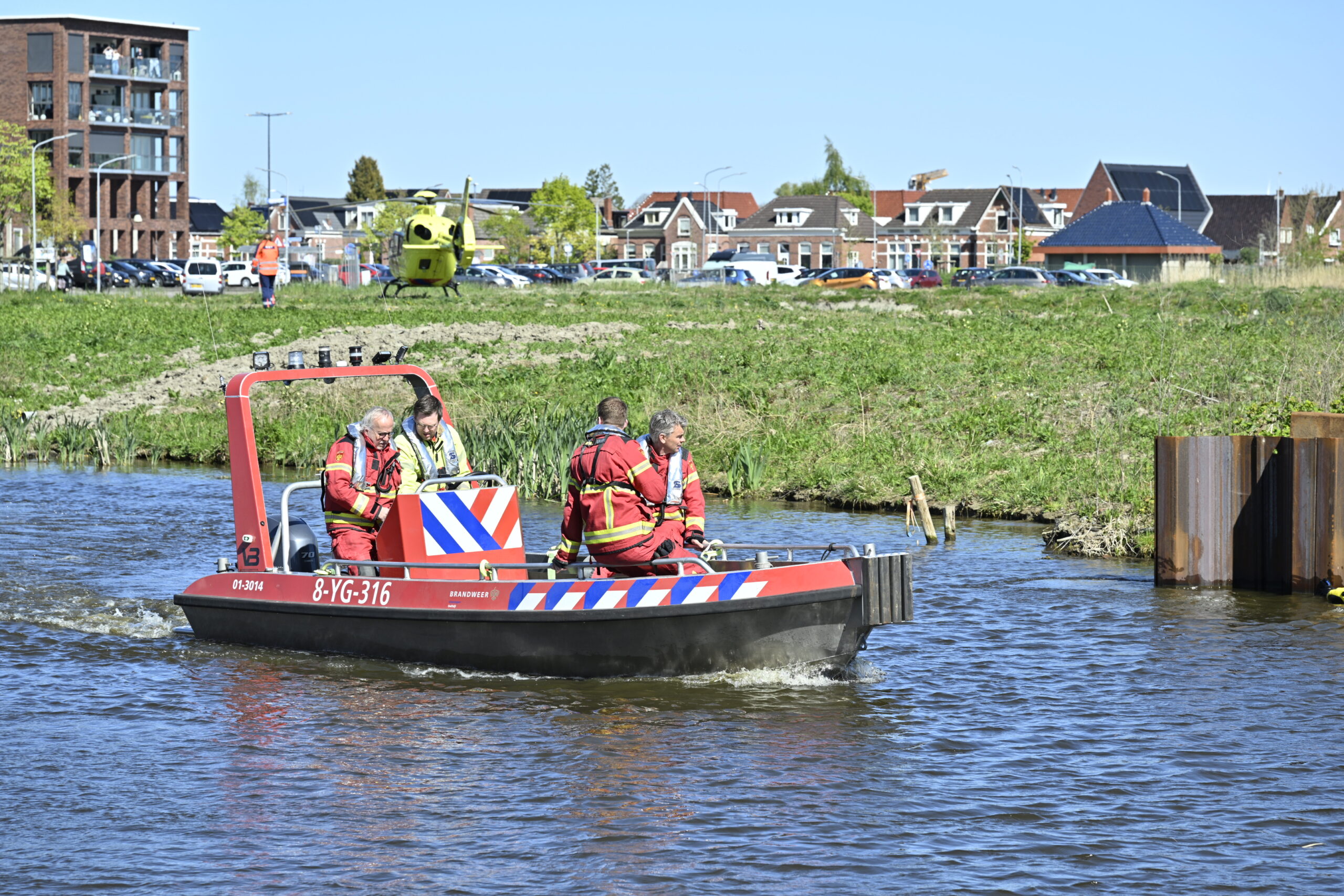 Red rescue boat with four crew members in life jackets on a river, with a yellow helicopter and urban buildings in the background on the shore.