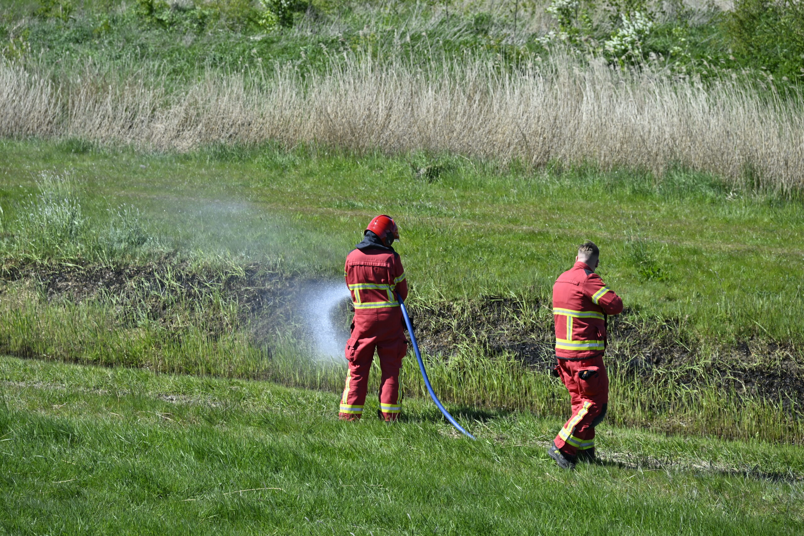 Two firefighters in red turnout gear spray water on a grassy ditch in a field.