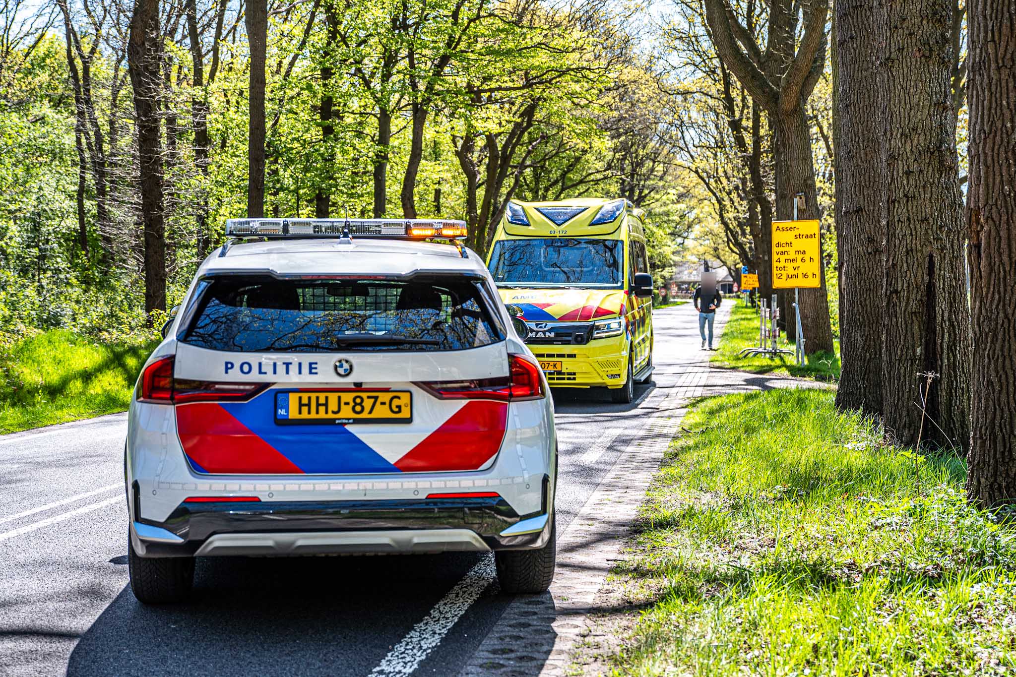 Police car with blue-red chevrons in the foreground and a yellow ambulance behind on a tree-lined street, suggesting an emergency scene.