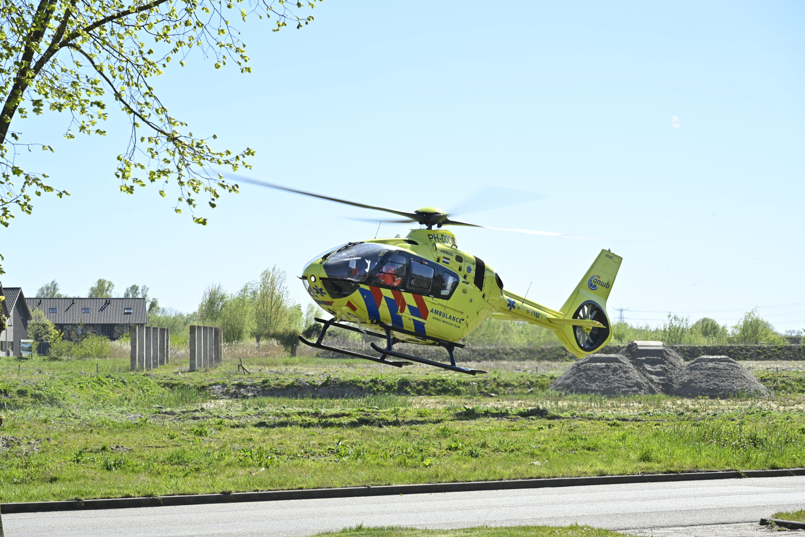 Yellow medical helicopter hovering low over a grassy field near a dirt mound, with houses and trees in the background during the day