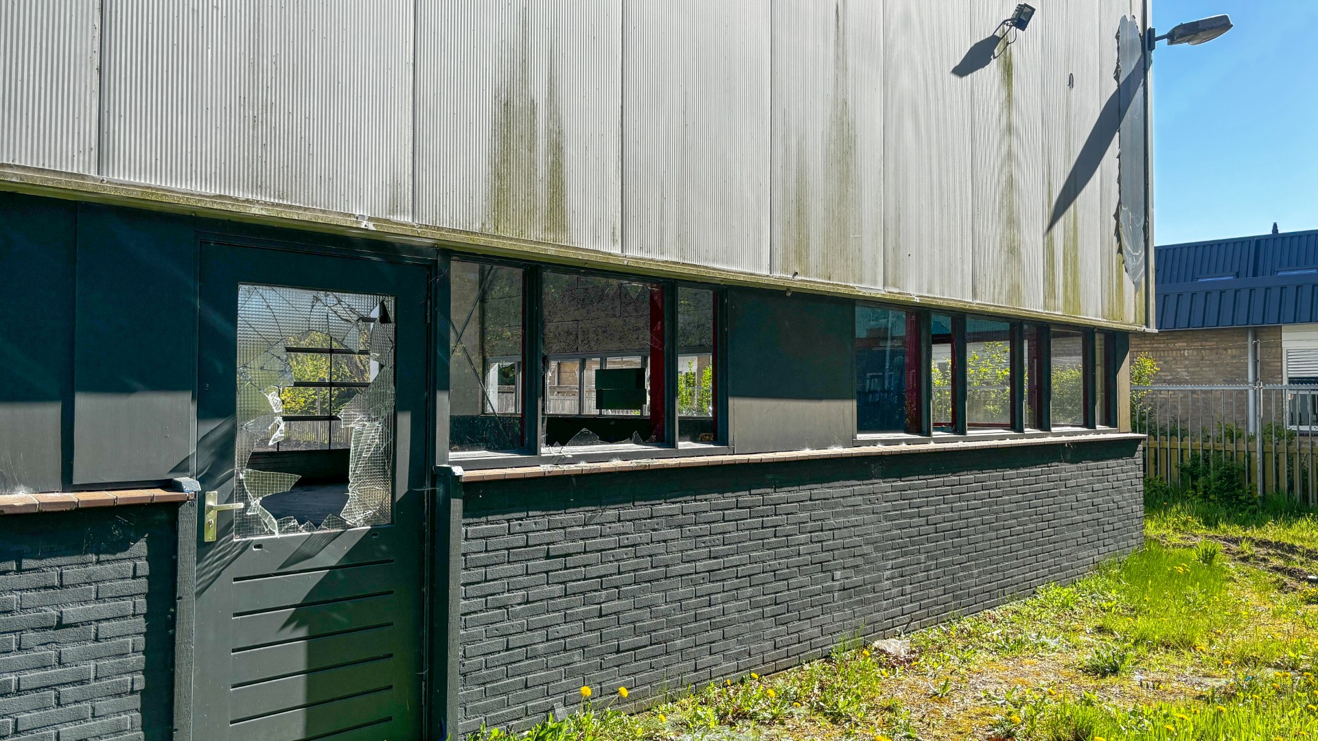 Abandoned storefront with a brick base, dark siding, and cracked glass on the door; overgrown lot in front under a corrugated-metal facade.