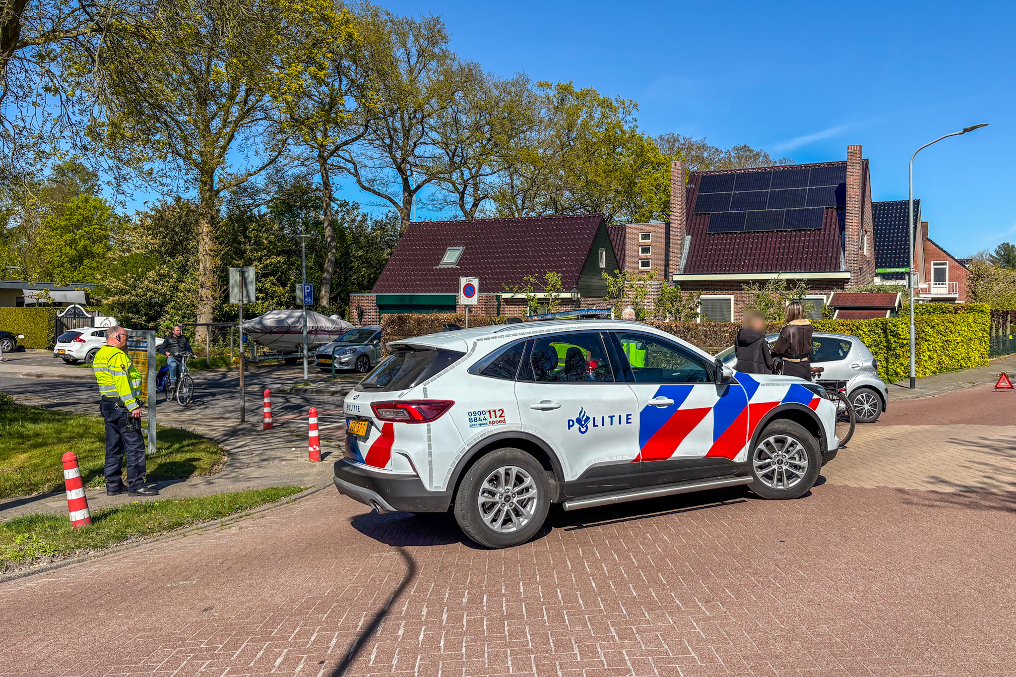 Dutch police car with blue-red chevron markings in foreground; officer in high-visibility jacket standing nearby on a residential street with bicycles and pedestrians.