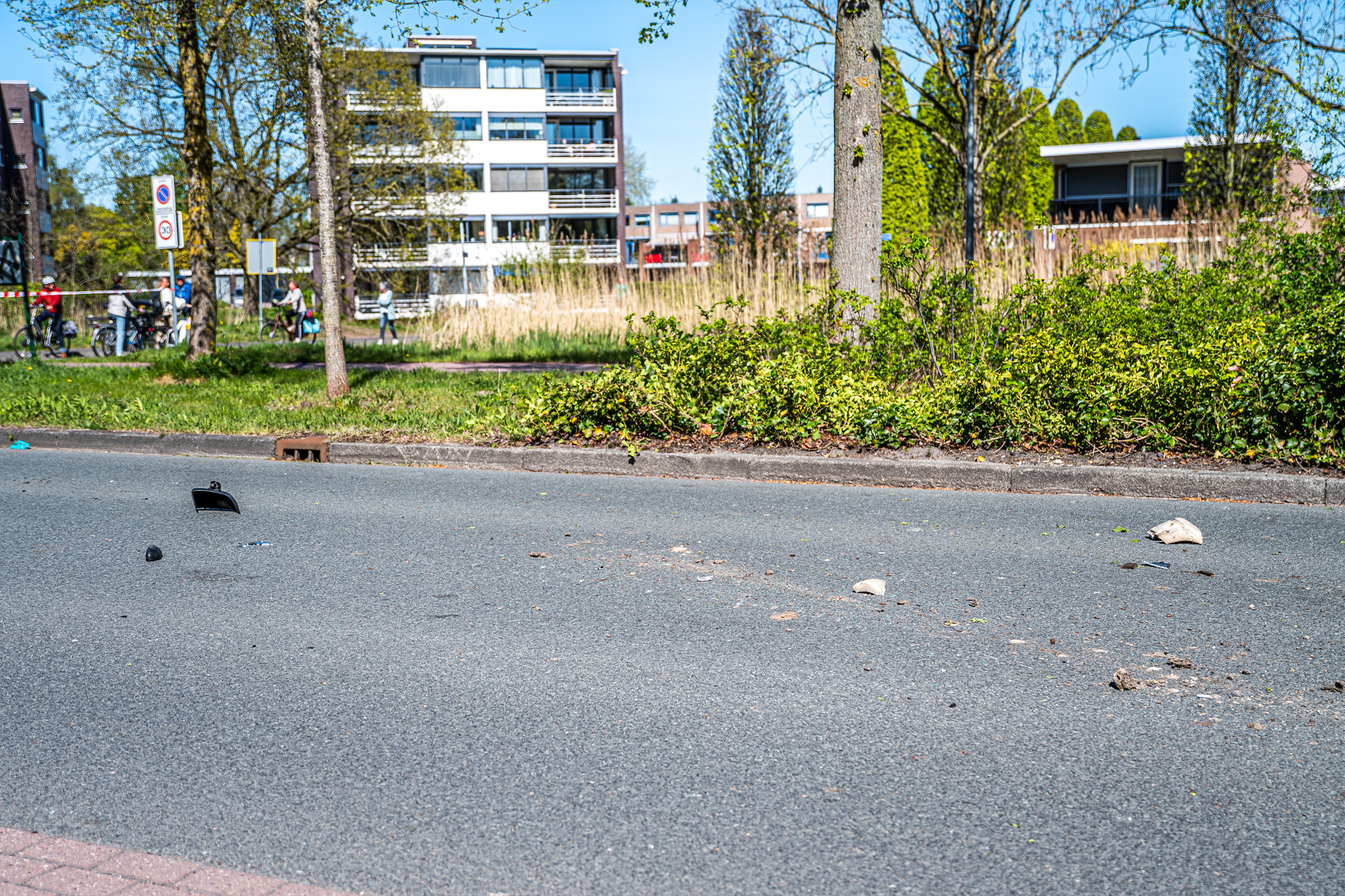 Urban street with scattered debris on the road as cyclists pass by in the background, with apartment buildings and trees behind them in daylight.