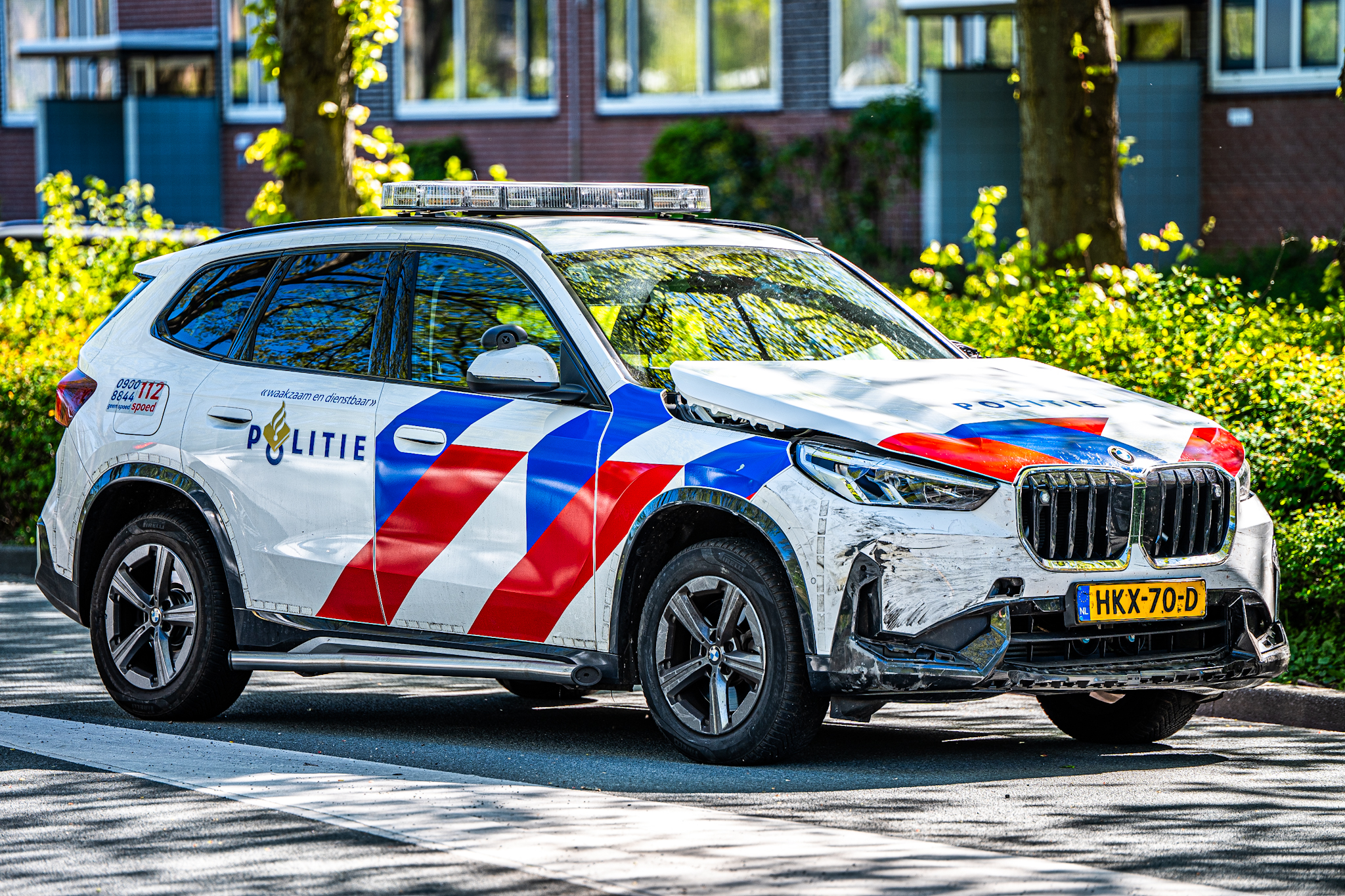 Dutch police SUV with roof emergency light bar, red–blue chevron stripes, and a damaged front end parked on a street in a residential area