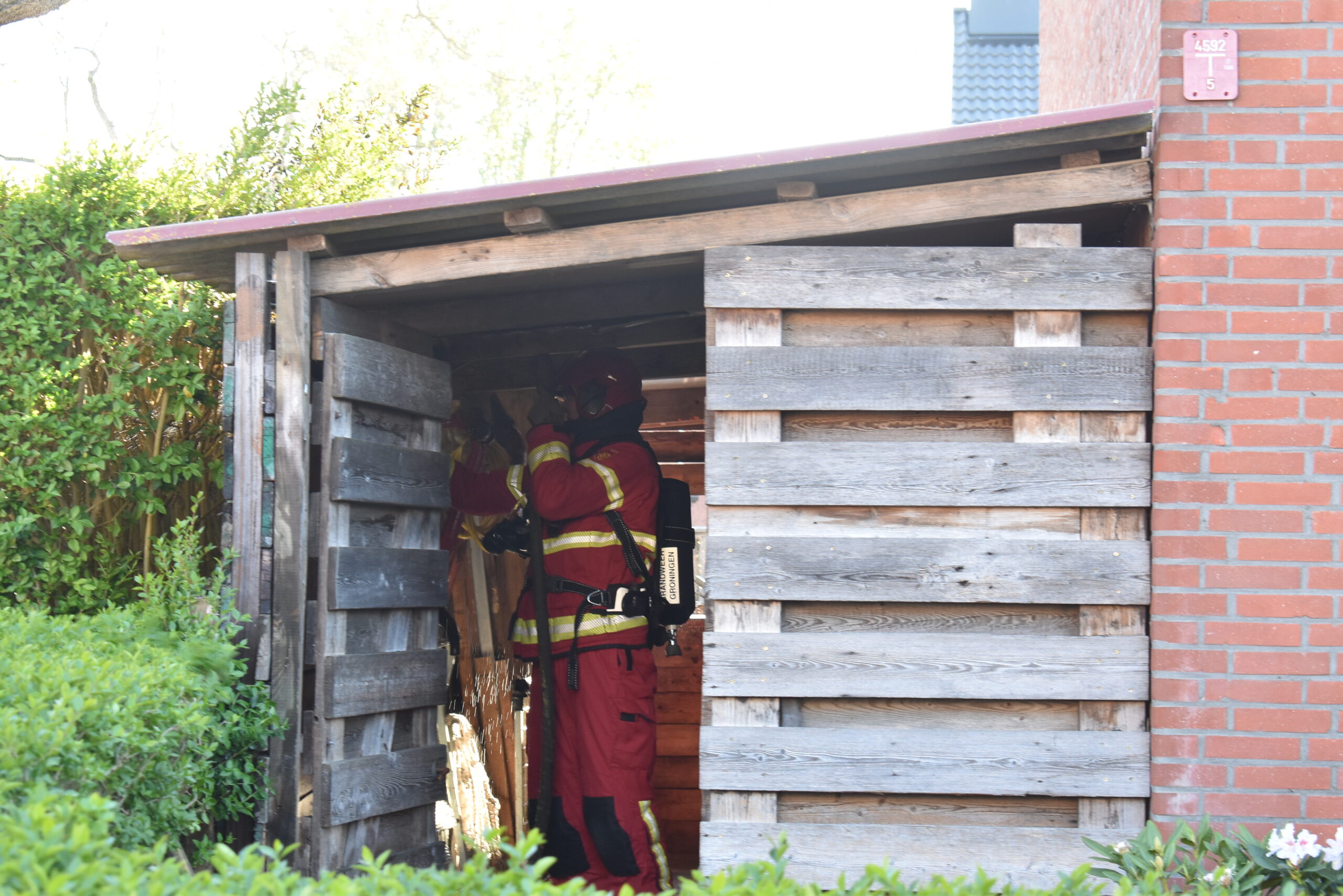 Firefighter in red turnout gear and breathing apparatus standing inside a wooden shed with a brick wall nearby, sparks visible from below.