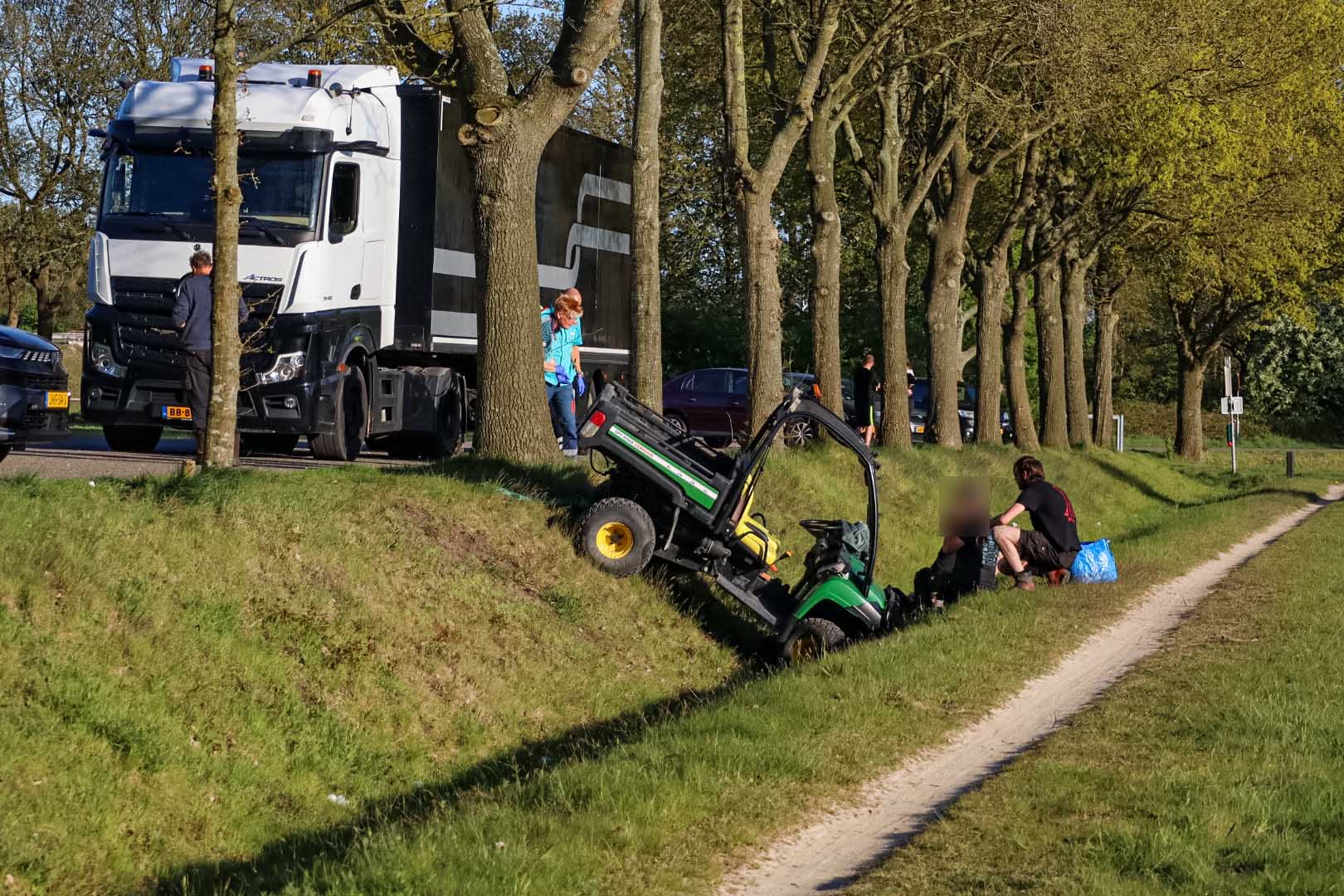 Large white semi-truck on a road beside trees; a green mini-loader is tipped in a ditch with people nearby.