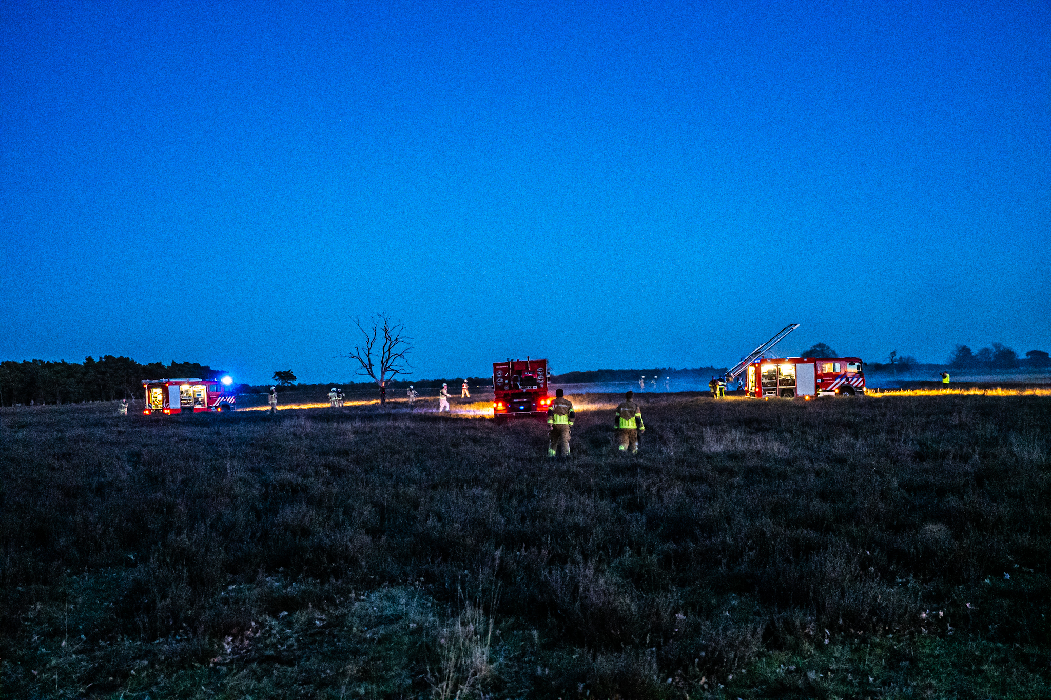 Firefighters and two fire trucks on a dark field at dusk, with one ladder extended and emergency lights visible.