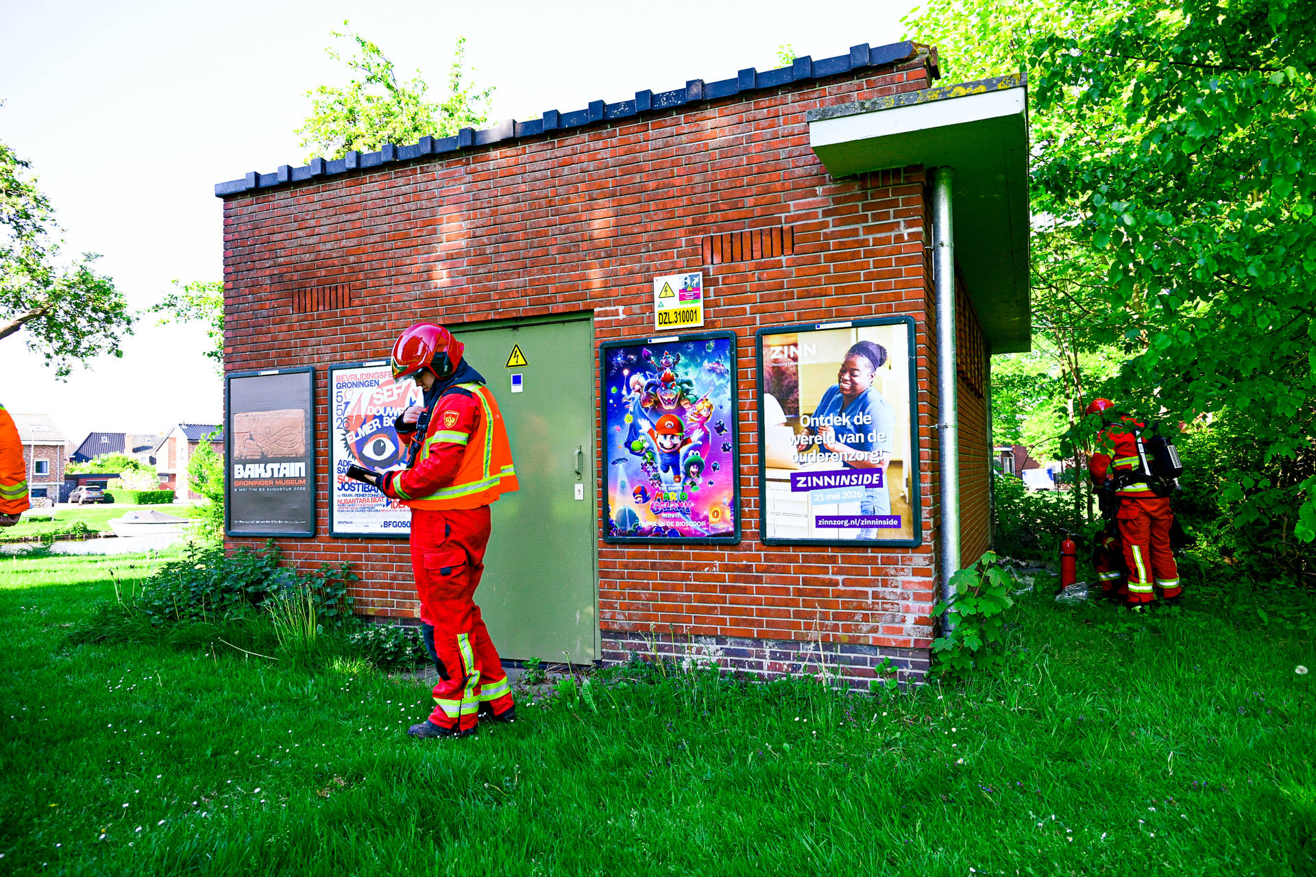 Firefighters in red gear stand outside a brick utility building with colorful posters on the wall.