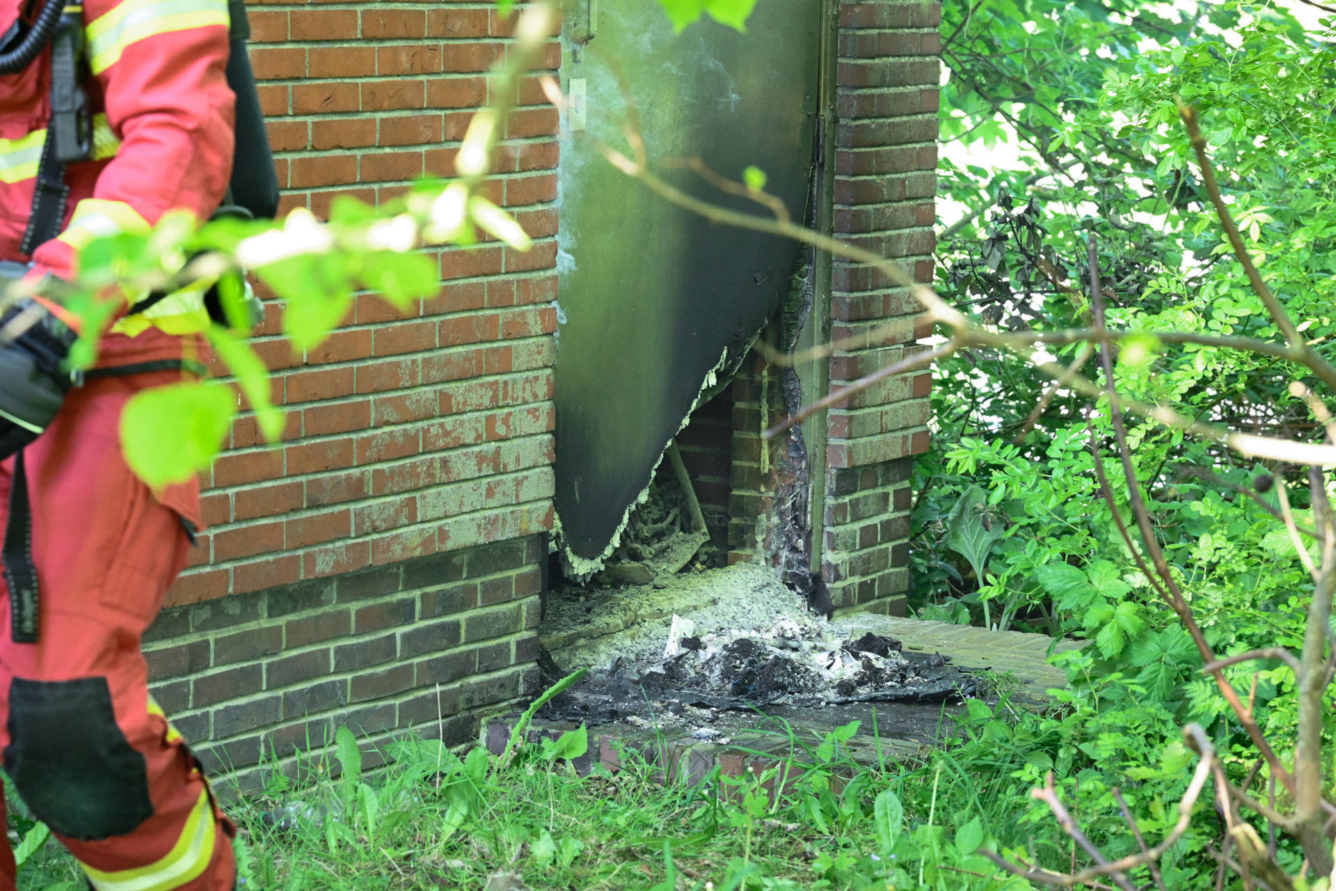 Firefighter in red turnout gear beside a damaged brick wall with a torn metal door and rubble on the ground, surrounded by overgrown plants.